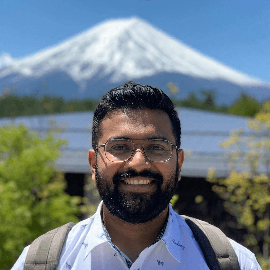 A smiling man with glasses and a beard stands before a majestic snow-capped mountain under a clear blue sky, embodying a.