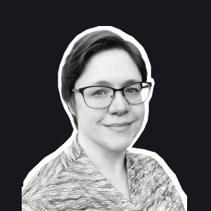 A smiling woman with short dark hair and glasses, wearing a patterned shirt, stands against a dark background embodying.
