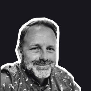 A smiling man with a beard, wearing a patterned shirt, set against a dark background embodying the friendly support.